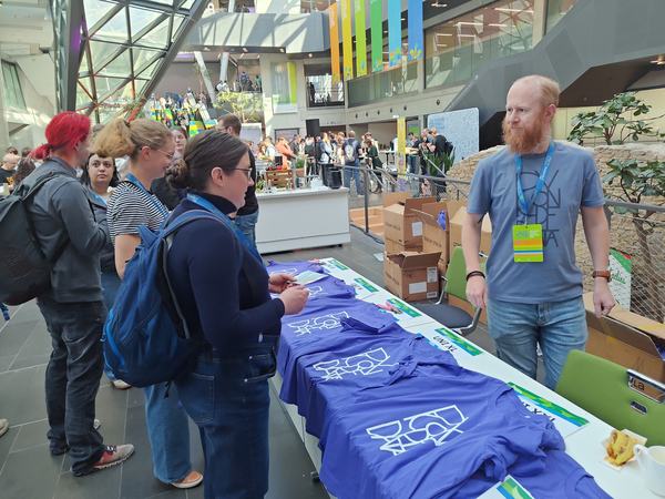 A photo of the EDC Lead Peter Holt volunteering at PyConDE & PyData in front of a table of conference t-shirts